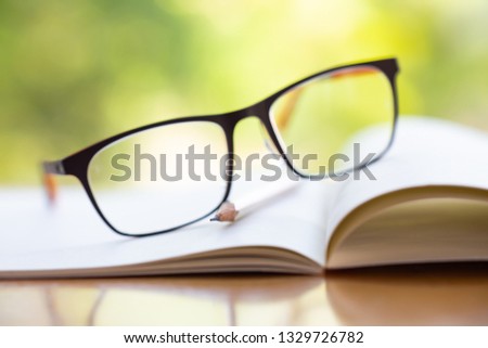 Black eyeglasses, white pencil with white notebook on wooden table, Bokeh garden background, Close up & Macro shot, Selective focus, Stationery concept