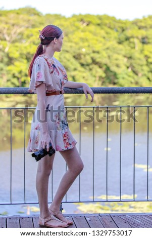 Young woman standing on banks of a lagoon holding a digital camera while leaning on a fence