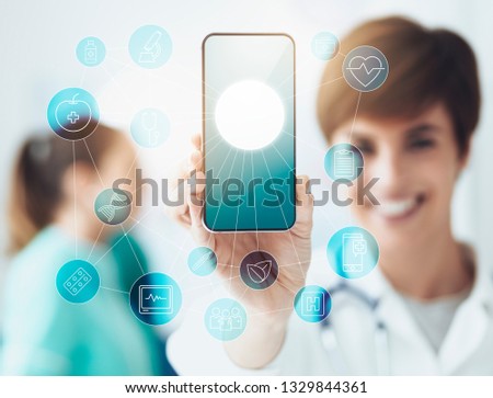 Smiling confident female doctor holding a touch screen smartphone with network of medical icons and healthcare app, medical staff working in the background: telemedicine and healthcare concept