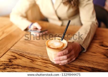 Close up of caucasian woman in jacket using smart phone and drinking coffee while sitting in cafeteria. Selective focus on hand with coffee.