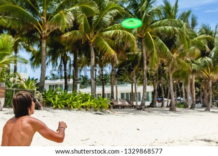 A man with long hair and sunglasses plays flying disc on a white sand beach. Green tall palm trees in the background.