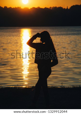 Silhouette of a young girl stands in fashion pose at the sunset on the river