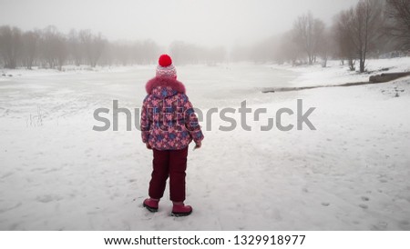 Winter day. The girl on the river bank looks at the lake. Fog and light rain with snow. Picture taken in Ukraine, Kiev region. Horizontal frame. Color image