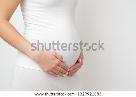 Woman in white clothes touching big belly with hand. Isolated on gray background. Emotional loving pregnancy time - 20 weeks. Baby expectation. Happiness and safety concept. Front side view.