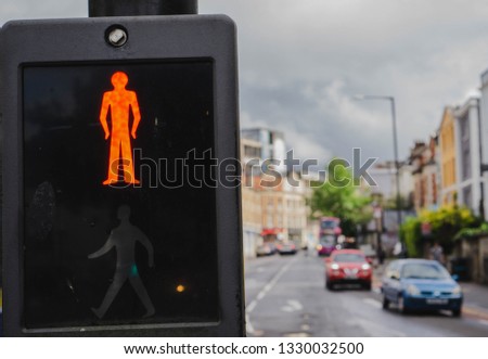 Close-up of pedestrian crossing signal in Bristol, England, showing the 'Red' man illuminated and lit up to stop people walking and crossing. Street building and passing cars in soft focus background.