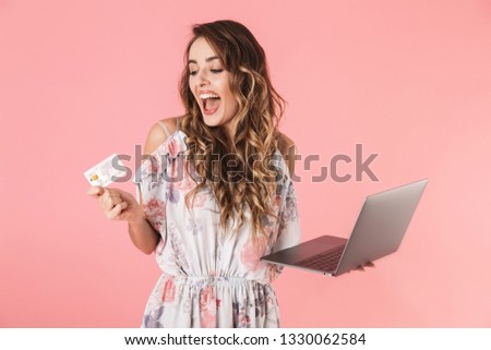 Photo of pleased woman 20s in dress holding silver laptop and credit card, isolated over pink background