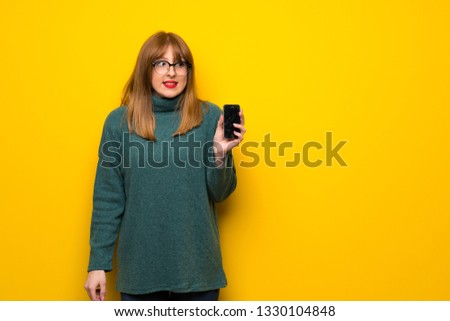 Woman with glasses over yellow wall with troubled holding broken smartphone