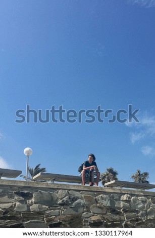 The young man sits on a bench with a skateboard and looks aside, against the background of the blue sky and palms. Dreamer, street racer. Teenage hobbies, board, rest.
