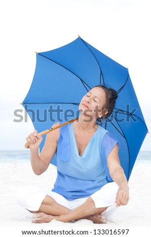 Portrait attractive mature woman sitting relaxed and happy with closed eyes and blue umbrella at beach, isolated with ocean and bright white sky as blurred background and copy space.
