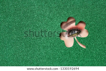 Conceptual photo of St. Patrick's Day. The Eye of the Young Beautiful Woman with Bright Golden Shadows and Expressive Eyebrows, Looks in the Shamrock Leaves Pattern out of Green Paper