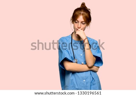 Young redhead nurse looking down with the hand on the chin over pink background