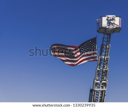 A photograph of an American Flag being Hoisted Up by a Fire Truck in Remembrance on September 11th, 2018