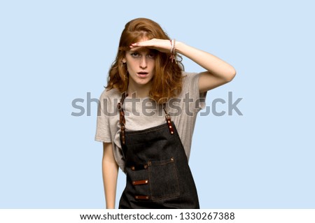 Young redhead woman with apron looking far away with hand to look something over blue background