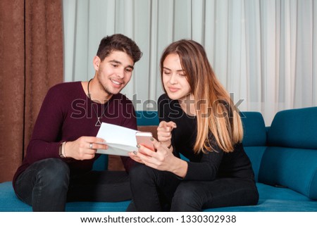 Young man and woman sharing smartphone while holding a tax or card statement paper and sitting on sofa at home. Calculating tax online concept.