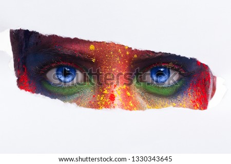 Girl with bright eyes make up looks through hole in white paper.
