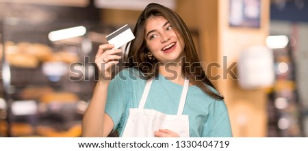 Girl with apron holding a credit card and thinking in a bakery