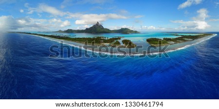 Aerial panoramic landscape view of the island of Bora Bora in French Polynesia with the Mont Otemanu mountain surrounded by a turquoise lagoon, motu atolls, reef barrier, and the South Pacific Ocean