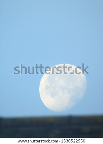 Detailed picture of the moon rising above the roof of a building during the day with blue sky.
