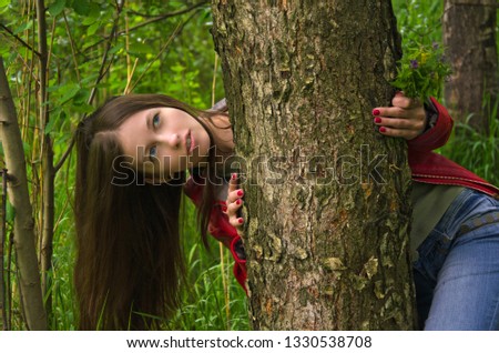 young girl with long black hair hiding behind a tree trunk in the woods

