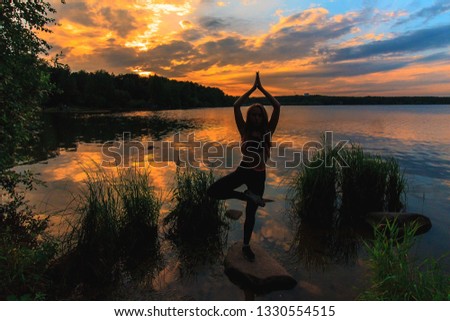 Woman standing in tree pose outdoors on a stone in the lake at sunset. Yoga nature concept.