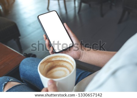 Mockup image of hands holding black mobile phone with blank screen while drinking coffee in modern cafe