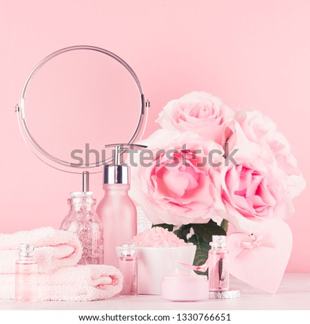 Romantic bathroom interior in pastel pink, silver color - flowers, round mirror, bath accessories, cosmetic products - cream, salt, rose oil, soap on white wood table.