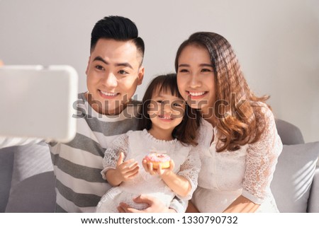 Beautiful young mother, father and their daughter are making selfie using a phone and smiling while sitting on the sofa at home