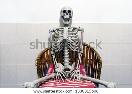 Fake human skeleton sitting on a chair in white room.