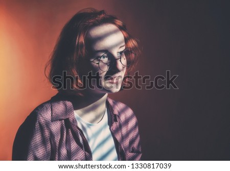 Red-haired girl with glasses in a plaid shirt and a white t-shirt is sad at the window with blinds