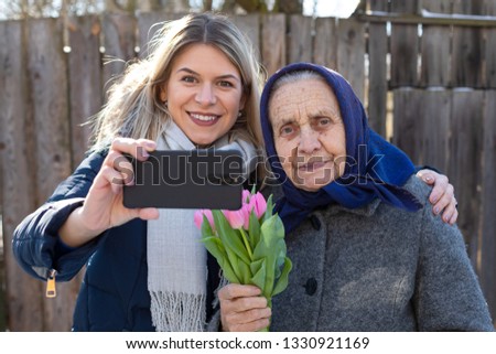 Picture of joyful women taking a selfie outdoor - young beautiful woman with old granny holding a smart phone and flower bouquet, smiling to the camera