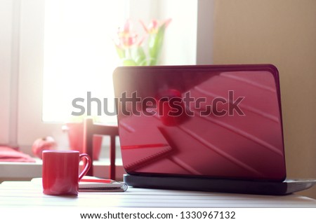 red cup of coffee and a notebook on the table are reflected in the lid of the laptop against the background of the window / day break