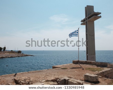 Stone cross and waving greek flag on the sea coast. Conon walls, Piraeus, Greece