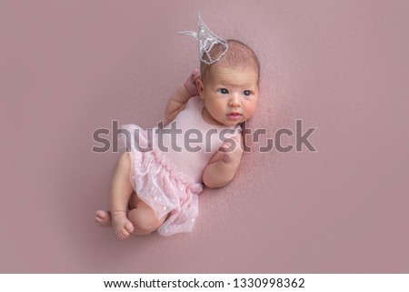 Caucasian (White) newborn baby girl in a pink polka-dot dress with ruffles on a pink background with a flowers bandage on her head. Piercing gaze. 13 days old. 