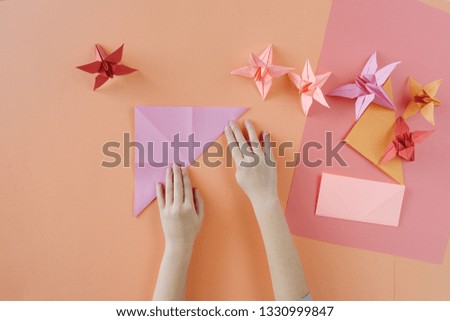 Children's hands do origami from colored paper on living coral background. Lesson of origami. Spring paper flower lie on a table. Top view, Flat lay style