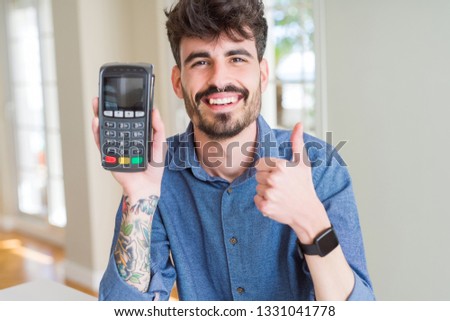 Young man holding dataphone point of sale as payment happy with big smile doing ok sign, thumb up with fingers, excellent sign