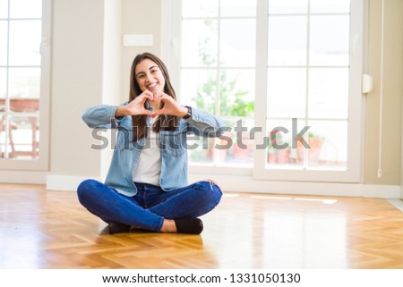 Beautiful young woman sitting on the floor at home smiling in love showing heart symbol and shape with hands. Romantic concept.