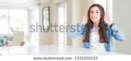 Wide angle picture of beautiful young woman sitting on white table at home Trying to hear both hands on ear gesture, curious for gossip. Hearing problem, deaf