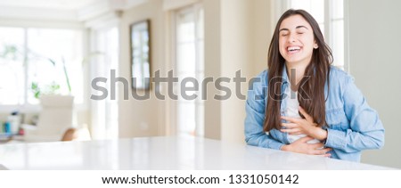 Wide angle picture of beautiful young woman sitting on white table at home Smiling and laughing hard out loud because funny crazy joke. Happy expression.