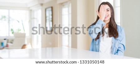 Wide angle picture of beautiful young woman sitting on white table at home covering one eye with hand with confident smile on face and surprise emotion.