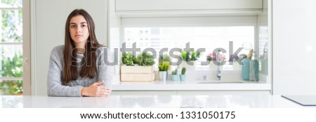 Wide angle picture of beautiful young woman sitting on white table at home with serious expression on face. Simple and natural looking at the camera.