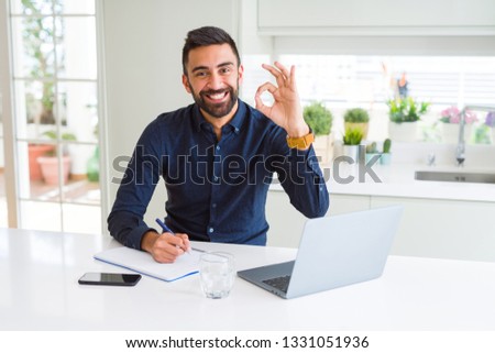Handsome hispanic man working using computer and writing on a paper doing ok sign with fingers, excellent symbol