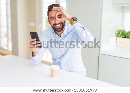 Handsome hispanic business man drinking coffee and using smartphone with happy face smiling doing ok sign with hand on eye looking through fingers