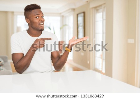 Handsome african american man on white table at home amazed and smiling to the camera while presenting with hand and pointing with finger.