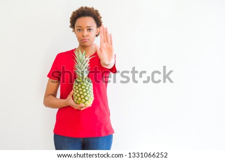 Young african american woman holding fresh healthy pineapple fruit with open hand doing stop sign with serious and confident expression, defense gesture