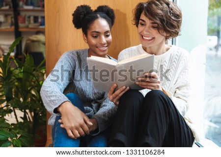 Two cheerful young girls students studying at library, reading a book together
