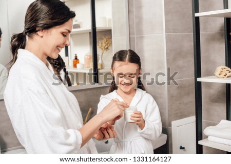 mother and daughter in white bathrobes with container of cosmetic cream in bathroom