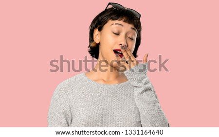Young woman with short hair yawning and covering wide open mouth with hand on isolated pink background