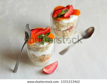 Two glasses of granola, yoghurt, tangerine, strawberry and mint leaves, spoons. Light background, horizontal orientation, copy space