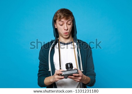 Portrait of amazed young man in casual clothes holding, looking on retro vintage photo camera isolated on blue wall background in studio. People sincere emotions lifestyle concept. Mock up copy space