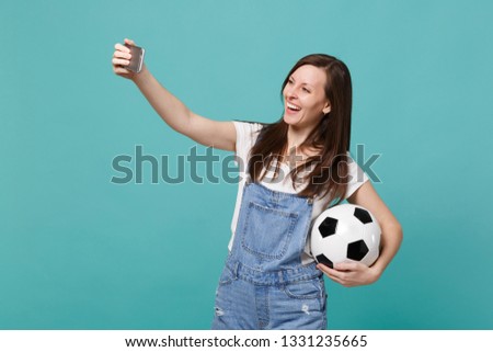 Smiling young woman football fan with soccer ball doing selfie shot on mobile phone isolated on blue turquoise background. People emotions, sport family leisure lifestyle concept. Mock up copy space